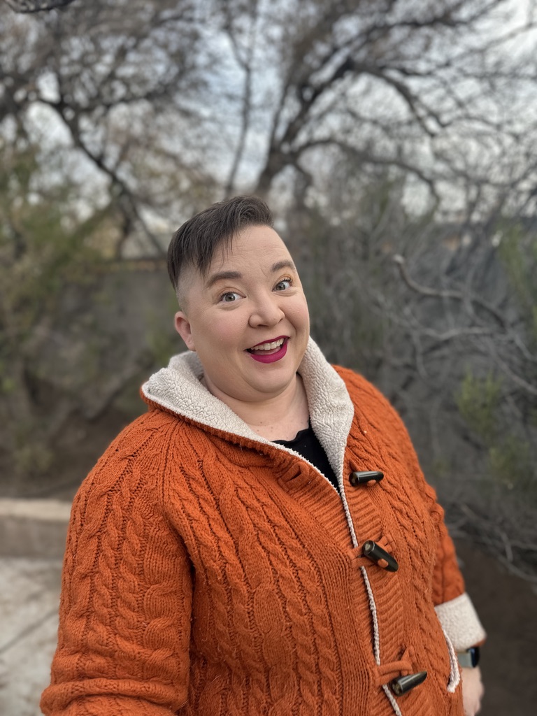 webs, smiling, outdoors in a rust cable-knit coat in front of bare winter trees.
